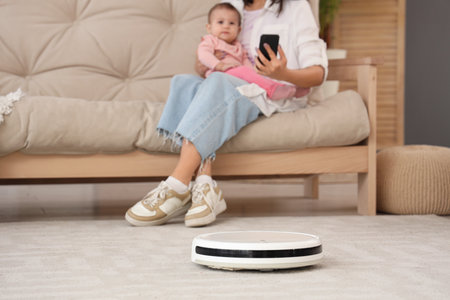 Mother with little baby and robot vacuum cleaner using mobile phone at home, closeupの写真素材