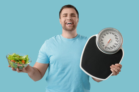 Young man with scales and salad on blue background. Weight loss conceptの写真素材