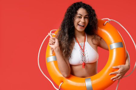 Beautiful young happy African-American female lifeguard with ring buoy pointing at something on red...