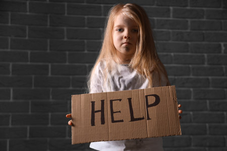 Homeless little girl holding piece of cardboard with word HELP near gray brick wallの写真素材