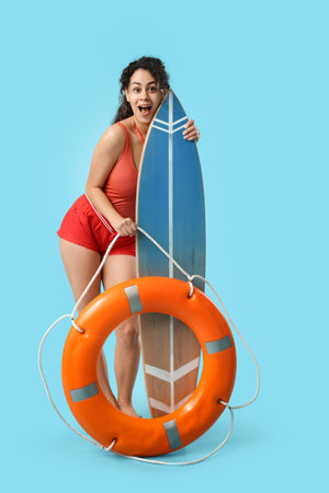 Beautiful young shocked African-American female lifeguard with surfboard and ring buoy on blue backgroundの写真素材