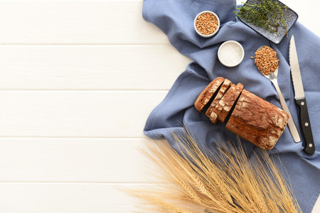Sliced loaf of bread, wheat spikelets, grains and thyme on white wooden background