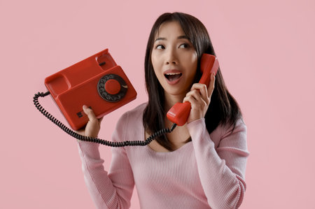 Young surprised Asian woman talking with retro phone on pink backgroundの写真素材