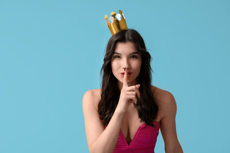 Beautiful young woman in stylish prom dress and paper crown showing silence gesture on blue backgroundの写真素材