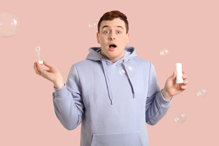 Young surprised man in hoodie with soap bubbles on pink background