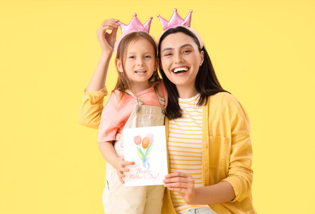 Young woman with her cute little daughter in crowns and greeting card on yellow background. Mother's...