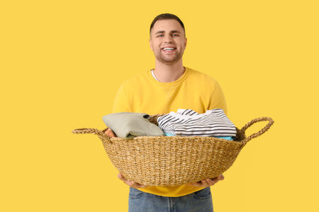 Young man holding basket with clothes on yellow backgroundの写真素材