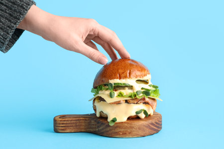 Female hand with tasty burger and wooden board on blue backgroundの写真素材