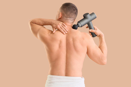 Muscular young man massaging his neck with percussive massager on beige background, back viewの写真素材