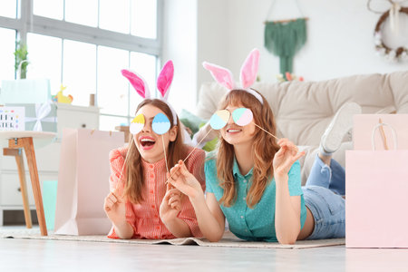 Happy young women in bunny ears headbands with paper shopping bags and decor for Easter lying on...