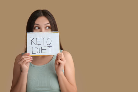 Beautiful young thoughtful Asian woman holding notebook with text KETO DIET on brown backgroundの写真素材
