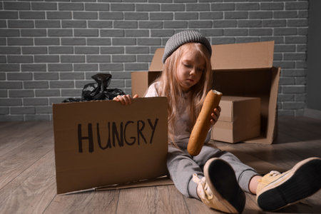 Homeless little girl with bread begging for food while sitting on floorの写真素材