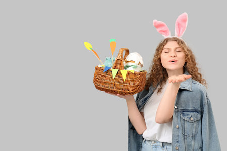 Beautiful young woman in bunny ears with Easter basket blowing kiss on gray background