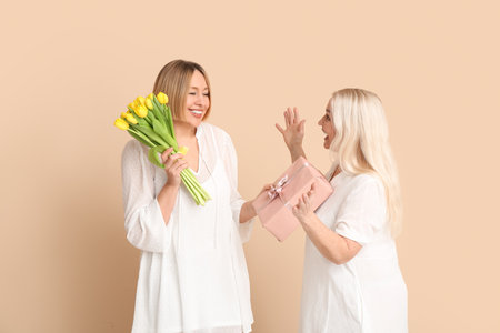 Adult woman greeting her mother with bouquet of tulips and gift box on beige background. International Women's Day celebrationの写真素材