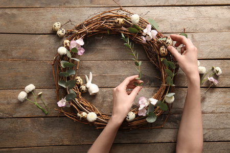 Woman making Easter wreath with quail eggs, flowers and plant branches on brown wooden backgroundの写真素材