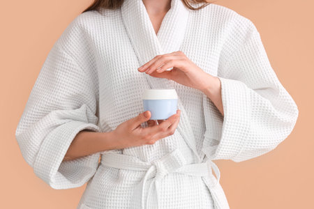 Woman in white bathrobe holding jar of cream on color background