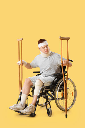 Injured young man after accident in wheelchair with crutches on yellow backgroundの写真素材