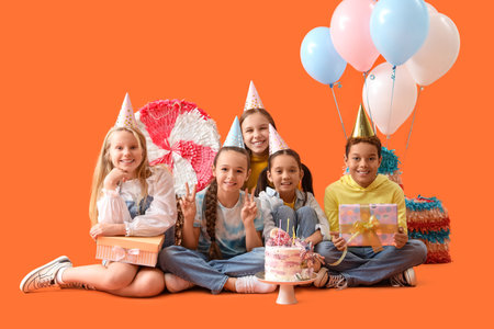 Cute little children in party hats with Birthday cake and gifts sitting on orange background