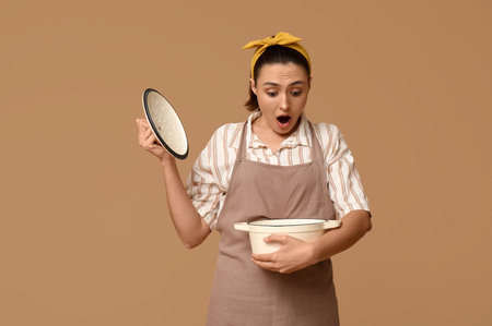 Portrait of shocked young housewife in apron with cooking pot on brown backgroundの写真素材
