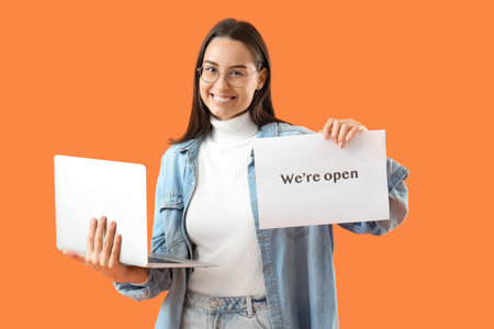 Female seller with OPEN sign and laptop on orange backgroundの写真素材