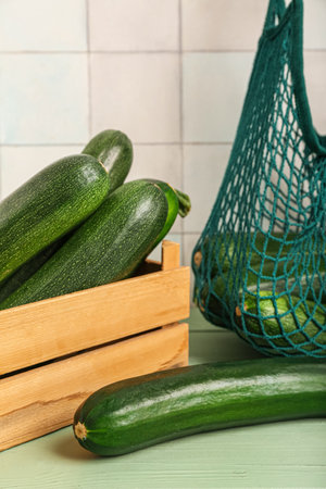Box and string bag with many fresh zucchini on green wooden table near white tile wallの写真素材