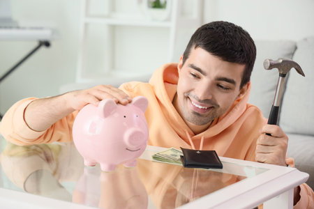Happy young man with hammer breaking piggy bank at homeの写真素材