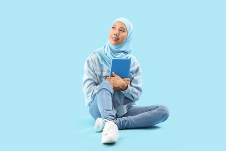 Beautiful young Asian woman in hijab with book sitting on blue backgroundの写真素材