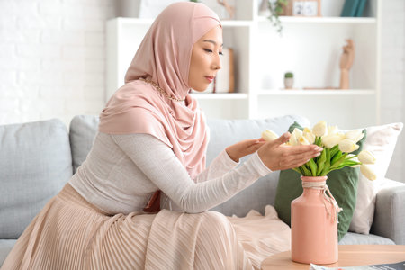 Beautiful young Asian woman in hijab with vase of flowers sitting on sofa at living roomの写真素材