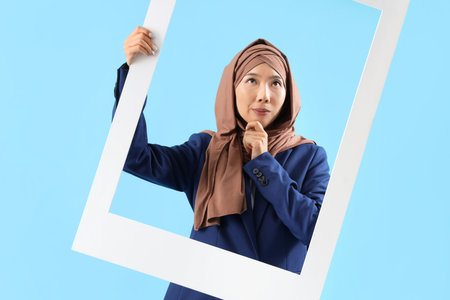 Thoughtful young Asian woman in hijab with frame on blue background