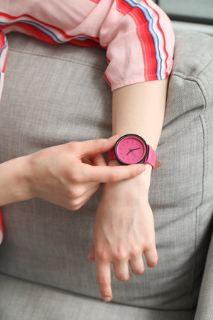 Young woman sitting on sofa and looking at stylish wristwatch in room, closeup