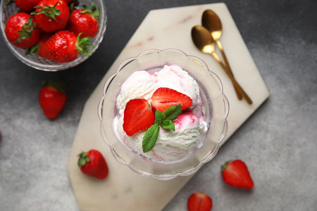 Bowl of vanilla ice cream with strawberries, mint and golden spoons on gray grunge table
