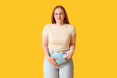 Young woman with hot water bottle having menstrual cramps on yellow background
