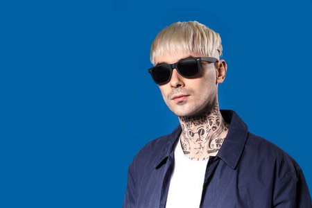 Young tattooed man in sunglasses on blue background, closeup