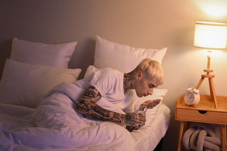 Young tattooed man with glass of water taking sleeping pill in bed at nightの写真素材