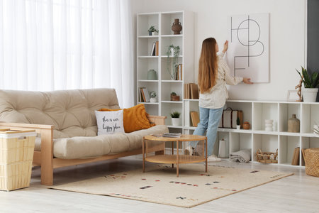 Young woman hanging poster on light wall in living room, back viewの写真素材