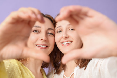 Beautiful mature woman with her daughter showing heart gesture on lilac background. Mother's Day celebrationの写真素材