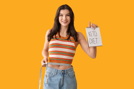 Happy smiling young woman holding notebook with text KETO DIET and measuring tape on orange backgroundの写真素材
