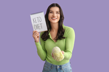 Happy smiling young woman holding notebook with text KETO DIET and cabbage on lilac backgroundの写真素材