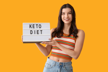 Happy smiling young woman holding board with text KETO DIET on orange backgroundの写真素材