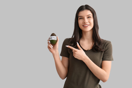 Happy smiling young woman pointing at avocado with the word KETO on white background. Diet concept