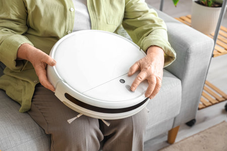Senior woman with robot vacuum cleaner sitting on sofa at homeの写真素材
