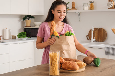 Beautiful young Asian woman with bread and paper bag full of fresh products in kitchenの写真素材