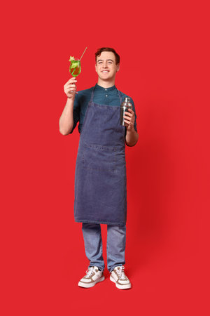 Young male bartender with glass of cocktail and shaker on red backgroundの写真素材