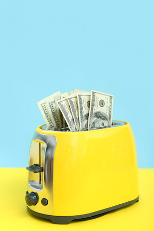Toaster with dollar banknotes on yellow table near blue wall