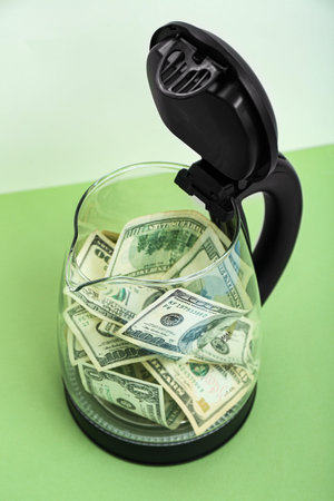 Electric pot with dollar banknotes on table near light wall
