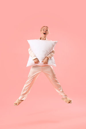 Young woman in pajamas with pillow jumping on pink backgroundの写真素材