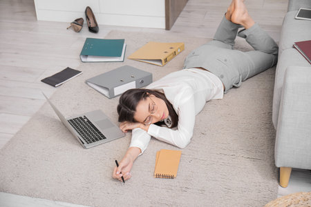 Tired young businesswoman sleeping on floor with laptop and notebook at homeの写真素材