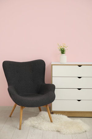 Dark gray armchair with dresser, fluffy rug and daisy near pink wall