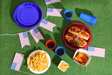 Tasty hot dogs with USA flags, cola and snacks on green grass background. American Independence Day...