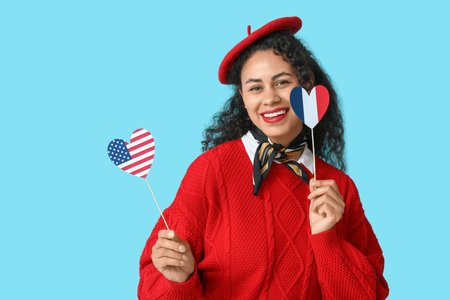Beautiful young happy African-American woman with USA and France flags in shape of hearts on blue backgroundの写真素材